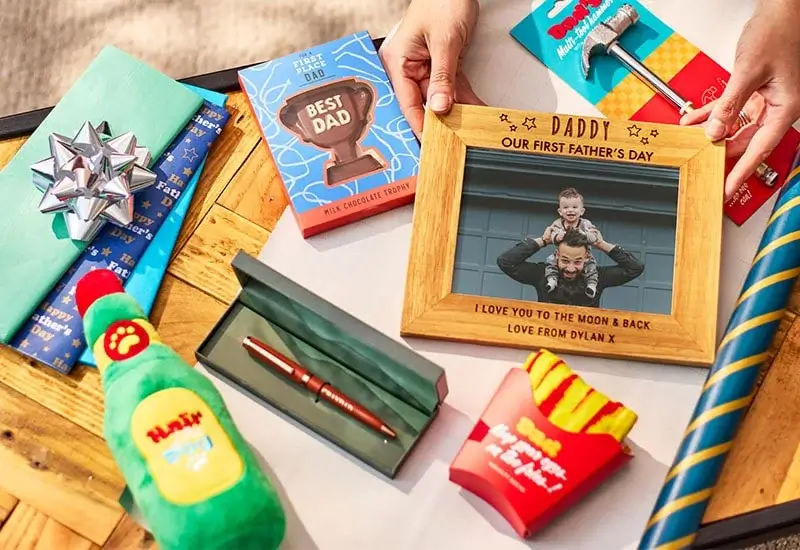 Father's Day gift selection on a wooden table, including a framed photo, chocolate trophy, wrapped presents, socks, and novelty items.